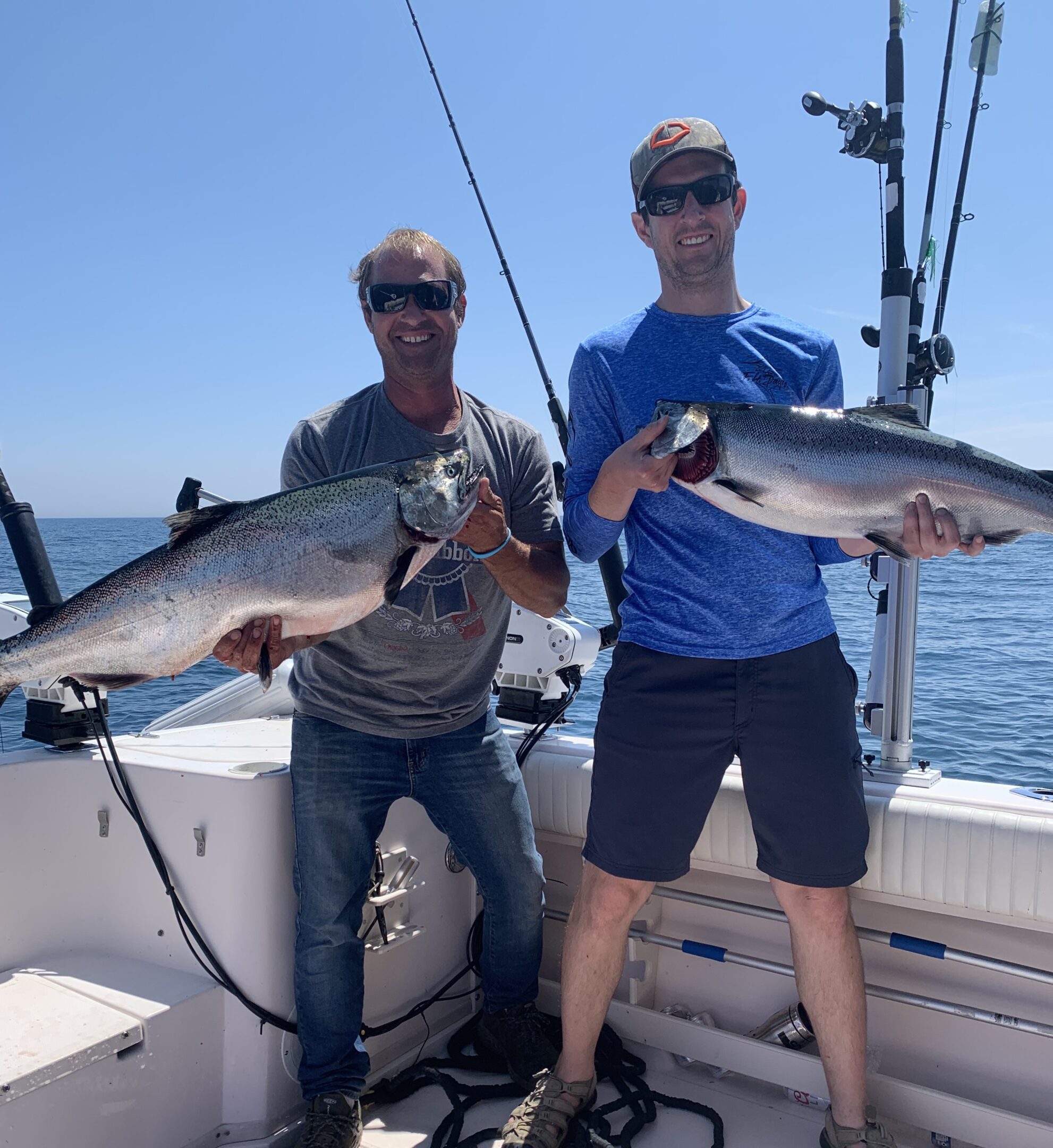 Two men holding large fish on a boat under clear blue sky.