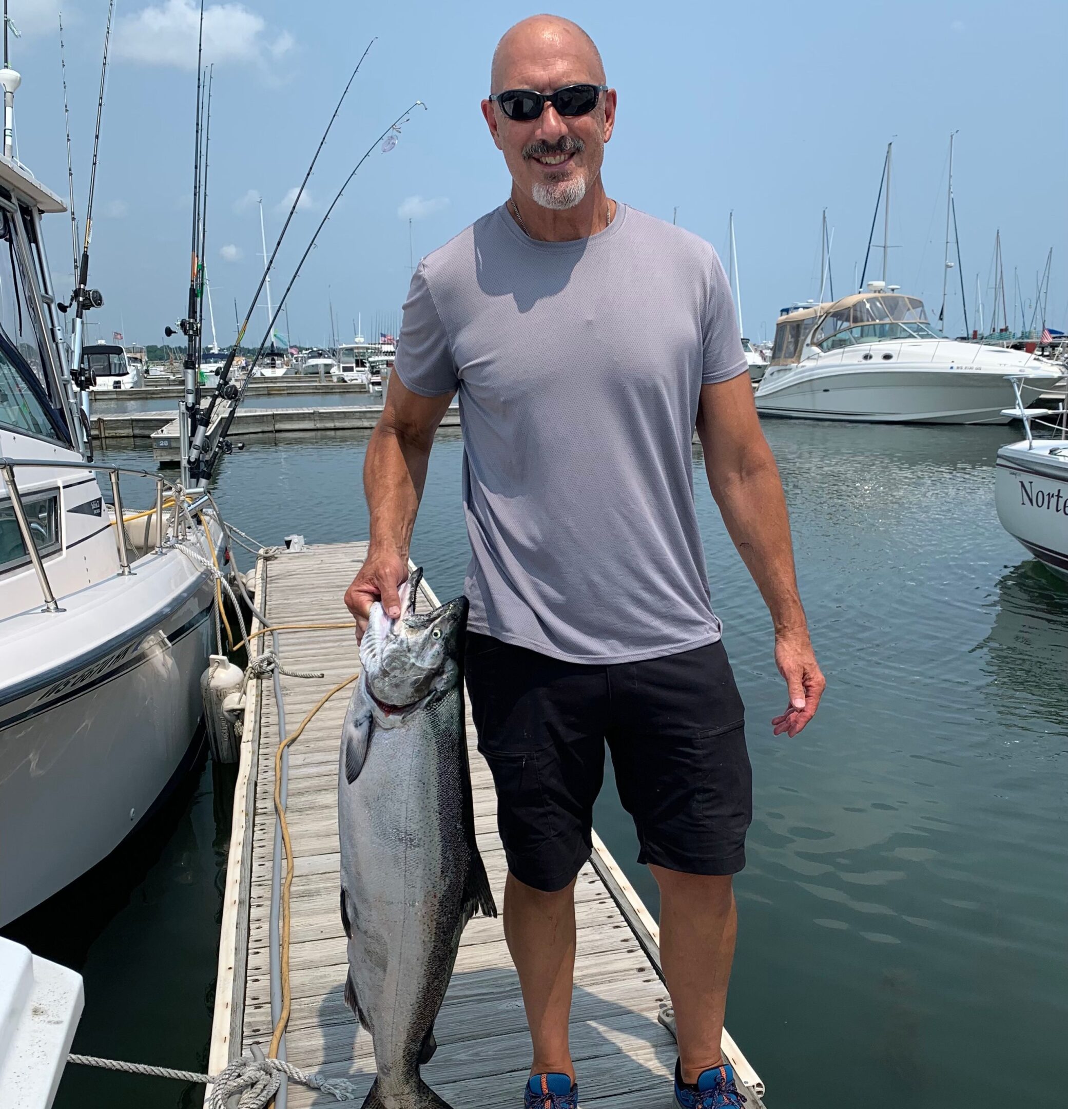 Man holding two large fish on a marina dock with boats in background.